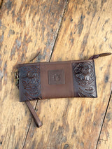Photo of front of dark brown leather clutch wallet with tooled leather on the sides and STS patch on front with wood back ground