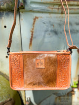 Close up image of the front of the Yipee Kiyay Claire crossbody purse. The purse has a brown cowhide panel in the middle with floral tooled veggie tan leather on each side and a leather patch in the middle with the STS brand.